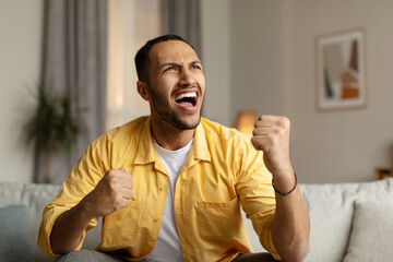 Emotional young black man shouting, gesturing YES in joy, celebrating victory, sitting on sofa at...