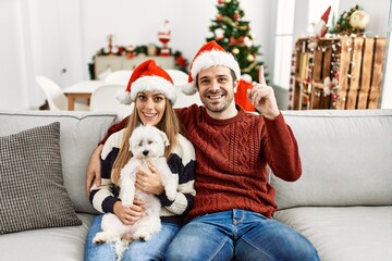 Young couple of wife and husband sitting on the sofa wearing christmas hat at home surprised with an idea or question pointing finger with happy face, number one