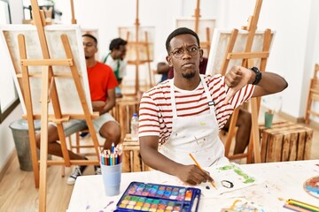 Young african man at art studio with angry face, negative sign showing dislike with thumbs down,...