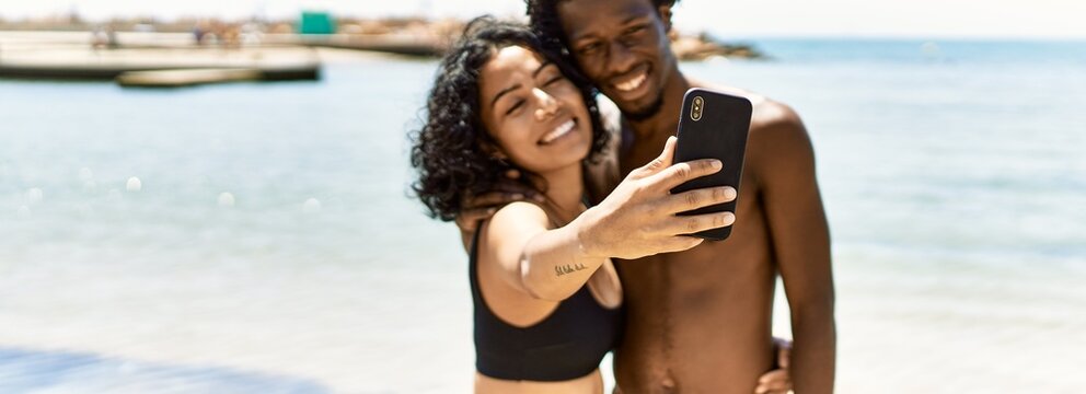 Young interracial tourist couple wearing swimwear making selfie by the smartphone at the beach.