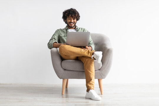 Joyful young indian sitting in armchair, chilling with modern laptop