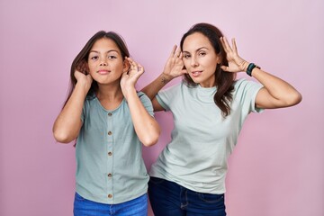 Young mother and daughter standing over pink background trying to hear both hands on ear gesture, curious for gossip. hearing problem, deaf