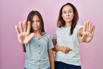 Young mother and daughter standing over pink background doing stop sing with palm of the hand....