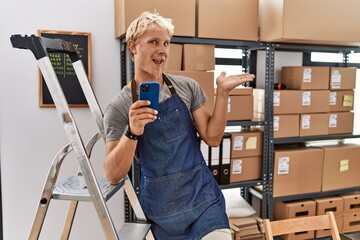 Young blond man using smartphone working at storehouse pointing aside with hands open palms showing...