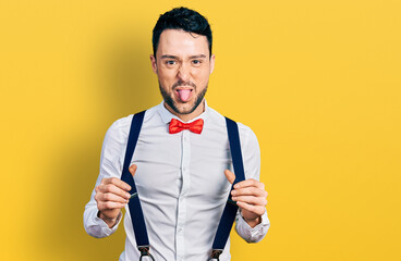 Hispanic man with beard wearing hipster look holding suspenders sticking tongue out happy with funny expression.