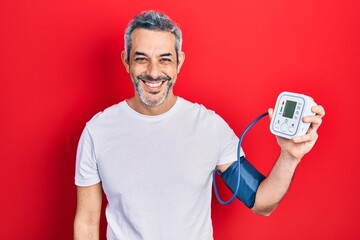Handsome middle age man with grey hair using blood pressure monitor looking positive and happy...