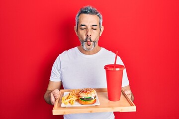 Handsome middle age man with grey hair eating a tasty classic burger with fries and soda making fish face with mouth and squinting eyes, crazy and comical.