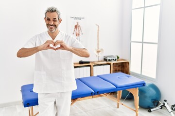 Middle age hispanic therapist man working at pain recovery clinic smiling in love doing heart symbol shape with hands. romantic concept.