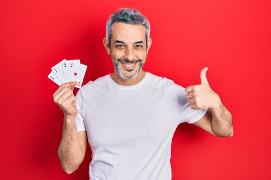 Handsome middle age man with grey hair holding poker cards smiling happy and positive, thumb up doing excellent and approval sign