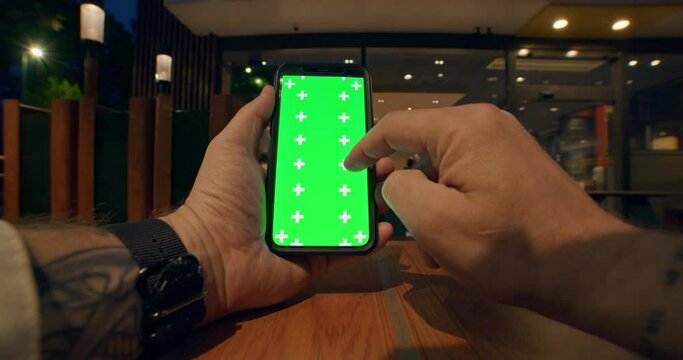 Close up view of young male tattooed hands with watches holding vertical and using mobile phone with green screen, scrolling smartphone touchscreen on table with soft lights of cafe on background