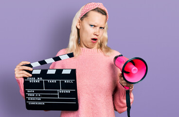 Young caucasian woman holding video film clapboard and megaphone in shock face, looking skeptical...