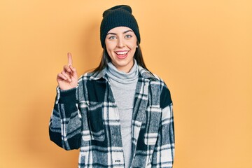 Beautiful woman with blue eyes wearing wool cap smiling with an idea or question pointing finger up with happy face, number one