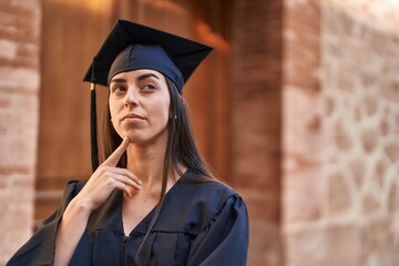 Young hispanic woman wearing graduated uniform standing with relaxed expression at university