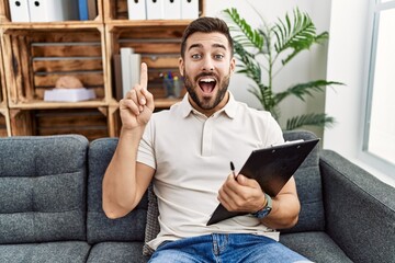 Handsome hispanic man holding clipboard working at psychology clinic smiling amazed and surprised and pointing up with fingers and raised arms.