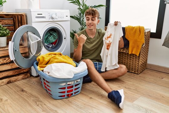 Young caucasian man doing laundry holding t shirt with stain pointing thumb up to the side smiling happy with open mouth