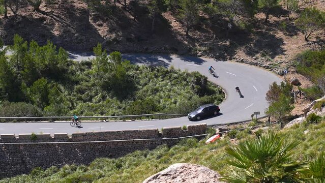 Athletes cycling on winding mountain road. Cyclists riding bicycles and car moving amidst trees. High angle view of bikers training during sunny day on the island of Mallorca.
