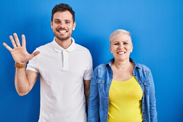 Young brazilian mother and son standing over blue background showing and pointing up with fingers number five while smiling confident and happy.