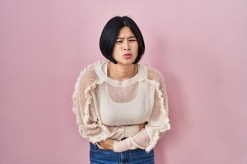 Fototapeta premium Young asian woman standing over pink background with hand on stomach because nausea, painful disease feeling unwell. ache concept.