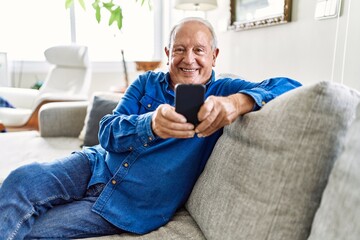 Senior man with grey hair sitting on the sofa at the living room of his house using smartphone