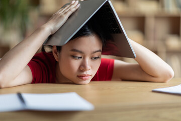 Tired frustrated teen asian girl makes roof with book on her head in library or room interior