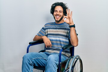 Handsome hispanic man sitting on wheelchair smiling positive doing ok sign with hand and fingers....