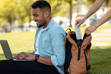 Female thief stealing smartphone from behind male bag, guy sitting on grass in park and working on...