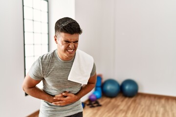 Young hispanic man wearing sportswear and towel at the gym with hand on stomach because nausea,...