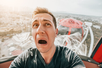 A guy with acrophobia or fear of heights screams with funny emotions on his face from the view from a high Ferris wheel in an amusement park - obrazy, fototapety, plakaty