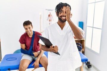 Obraz premium Young hispanic man working at pain recovery clinic with a man with broken arm covering one eye with hand, confident smile on face and surprise emotion.