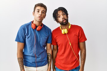 Young hispanic brothers standing over isolated background wearing headphones making fish face with...