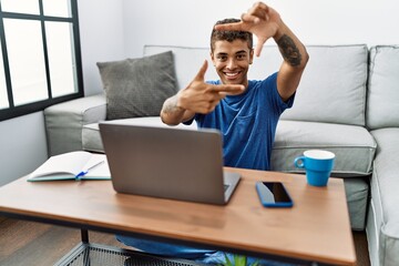 Young handsome hispanic man using laptop sitting on the floor smiling making frame with hands and fingers with happy face. creativity and photography concept.