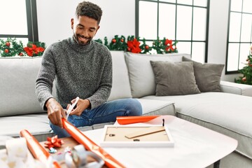 Young african american man smiling happy wrapping christmas gift sitting on the sofa at home. - obrazy, fototapety, plakaty