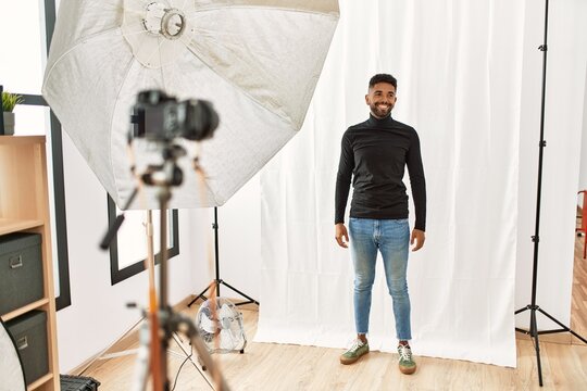 Young hispanic man with beard posing as model at photography studio looking away to side with smile on face, natural expression. laughing confident.