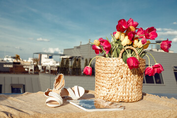 Composition of straw bag with bunch of pink, yellow flowers tulips, beige sandals and magazine on...