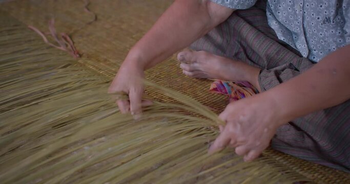 Cut in shot of an Asian female farmer who is an elderly native artisan weave mat from bamboo that are cut into strips for weaving folk crafts in rural areas of Thailand, southeast Asia.