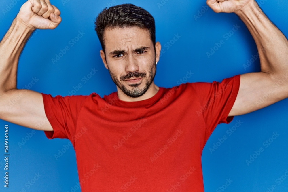 Wall mural young hispanic man with beard wearing red t shirt over blue background showing arms muscles smiling 