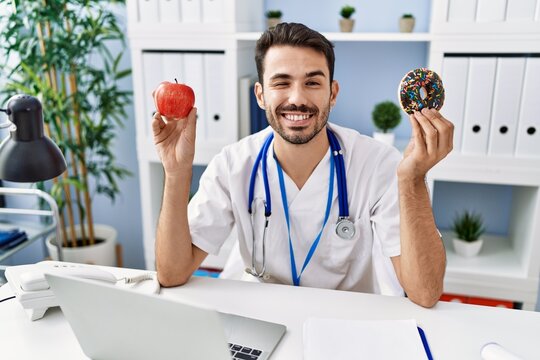 Young Hispanic Dietitian Man Holding Doughnut And Apple Winking Looking At The Camera With Sexy Expression, Cheerful And Happy Face.