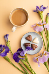 Fototapeta na wymiar Cup of cioffee with chocolate candies and lilac iris flowers on orange pastel background. top view, close up.