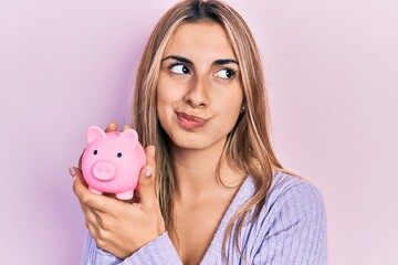 Beautiful hispanic woman holding piggy bank smiling looking to the side and staring away thinking.