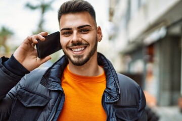 Handsome hispanic man with beard smiling happy and confident at the city wearing winter coat speaking on the phone