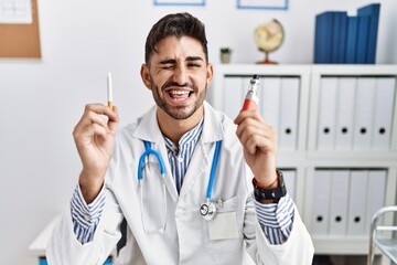 Young doctor man holding electronic cigarette at medical clinic winking looking at the camera with...