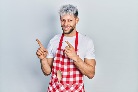 Young hispanic man with modern dyed hair wearing apron smiling and looking at the camera pointing with two hands and fingers to the side.