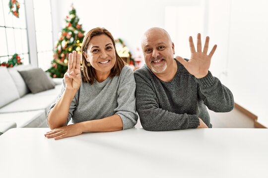 Middle age hispanic couple sitting on the table by christmas tree showing and pointing up with fingers number eight while smiling confident and happy.