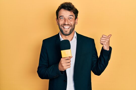 Handsome Man With Beard Holding Reporter Microphone Pointing Thumb Up To The Side Smiling Happy With Open Mouth