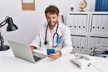 Young hispanic man wearing doctor uniform using smartphone at clinic