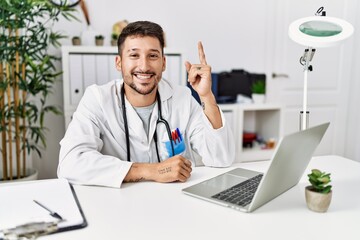 Young doctor working at the clinic using computer laptop pointing finger up with successful idea. exited and happy. number one.