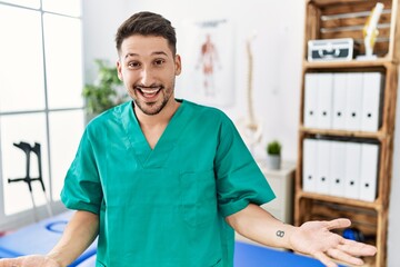 Young physiotherapist man working at pain recovery clinic smiling cheerful with open arms as friendly welcome, positive and confident greetings