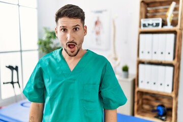 Young physiotherapist man working at pain recovery clinic in shock face, looking skeptical and sarcastic, surprised with open mouth