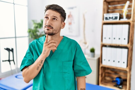 Young physiotherapist man working at pain recovery clinic thinking concentrated about doubt with finger on chin and looking up wondering