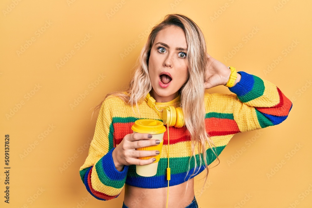 Sticker beautiful young blonde woman drinking cup of coffee wearing headphones crazy and scared with hands o